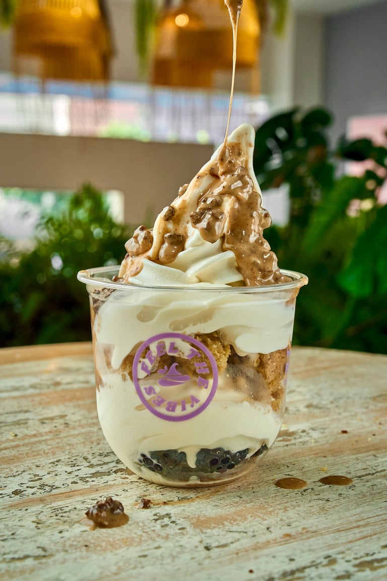Layered dessert with white cream, dark crumble base, and peanut butter swirl drizzle on top in a clear cup with purple branding label