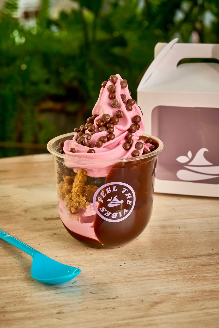 Pink frozen yogurt with chocolate pieces and cookie crumbles in a clear cup with blue spoon on wooden surface